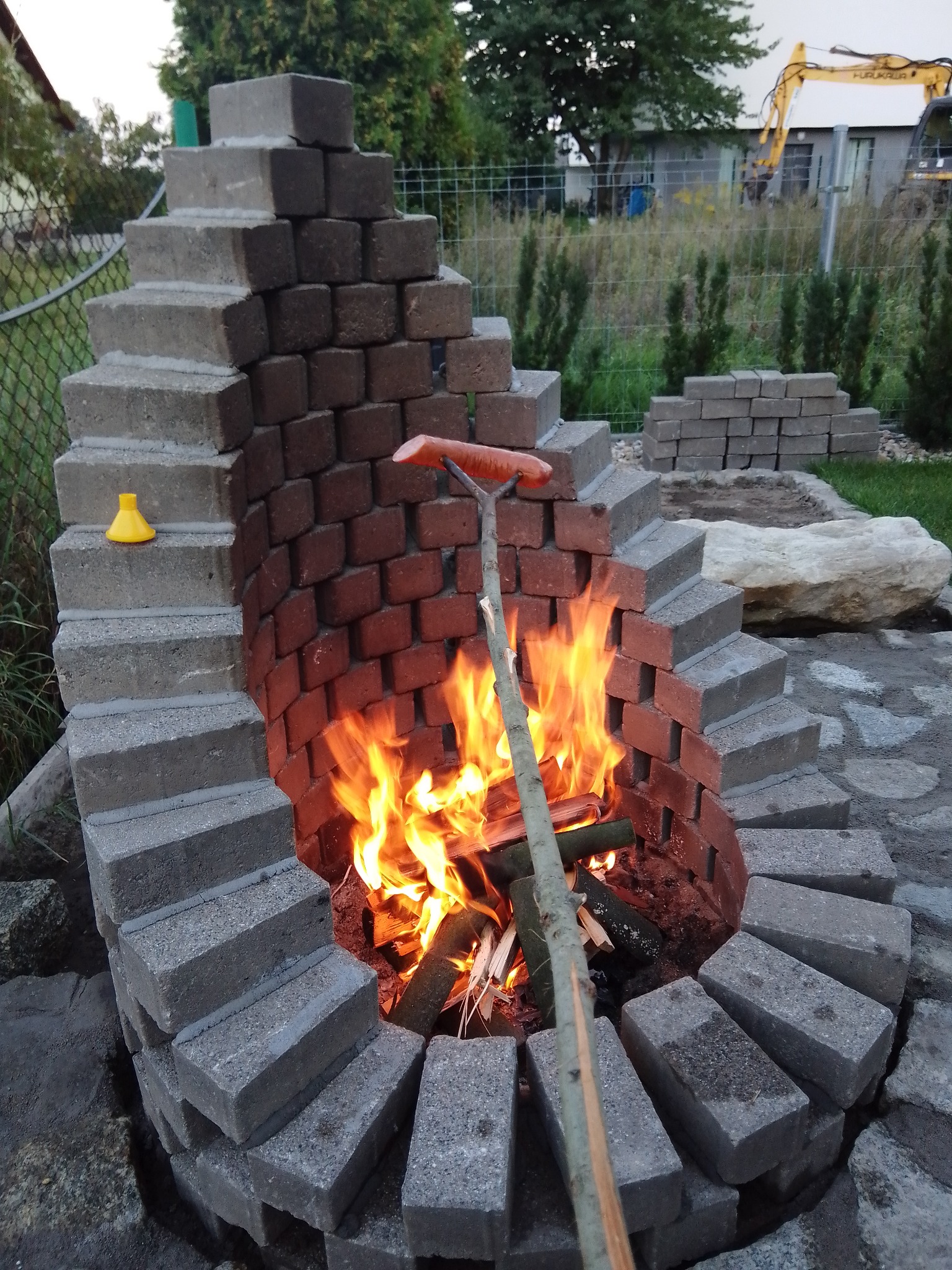 Ognisko z cegieł i betonowych bloczków, z płomieniami i rożnem z kiełbasą, w tle ogród i sprzęt budowlany.