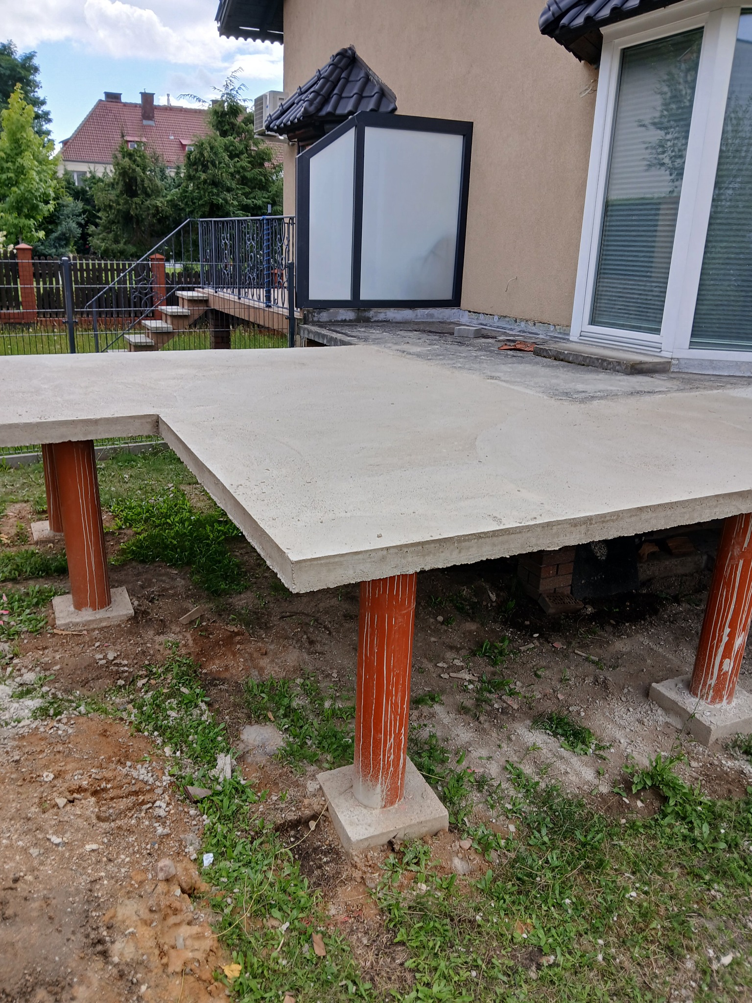 Przygotowany betonowy taras na solidnych, pomarańczowych podporach, z widocznym trawnikiem i fragmentem budynku w tle. Etap budowy tarasu.