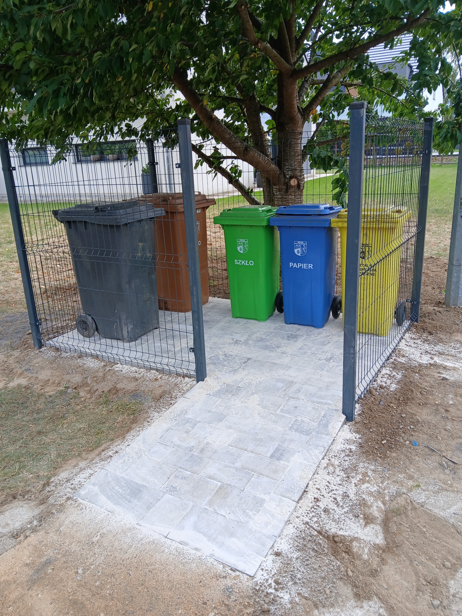 Wykonane z jasnej kostki dojście do zadaszonego, ogrodzonego miejsca na pojemniki do segregacji odpadów w kolorach: szarym, brązowym, zielonym, niebieskim i żółtym.