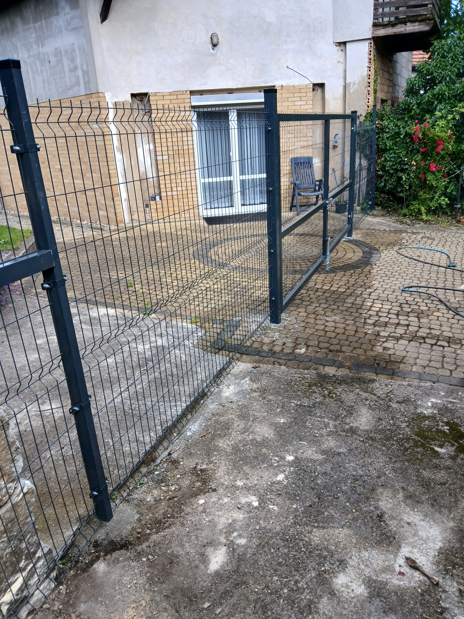 Nowoczesne ogrodzenie panelowe w kolorze grafitowym, oddzielające posesję od sąsiada. Widoczny fragment domu z oknem i fragment ogrodu z kostką brukową.