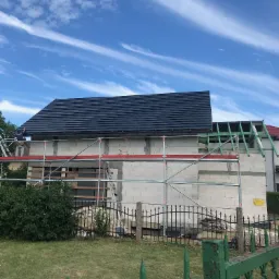 Budowa domu w trakcie, z częściowo położonym, ciemnym dachem i rusztowaniem, widok zza ogrodzenia.