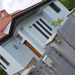 Damian Prauze - Jasnoszary dom jednorodzinny z numerem 55, widziany z dołu pod kątem. Czerwona dachówka, drewniane drzwi, szare ogrodzenie i skrzynki pocztowe.