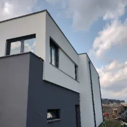Nowoczesny dom z dwukolorową elewacją: szary dół i biała góra, widoczne okna z czarnymi ramami, niebo z chmurami w tle, teren wokół domu częściowo zagospodarowany.