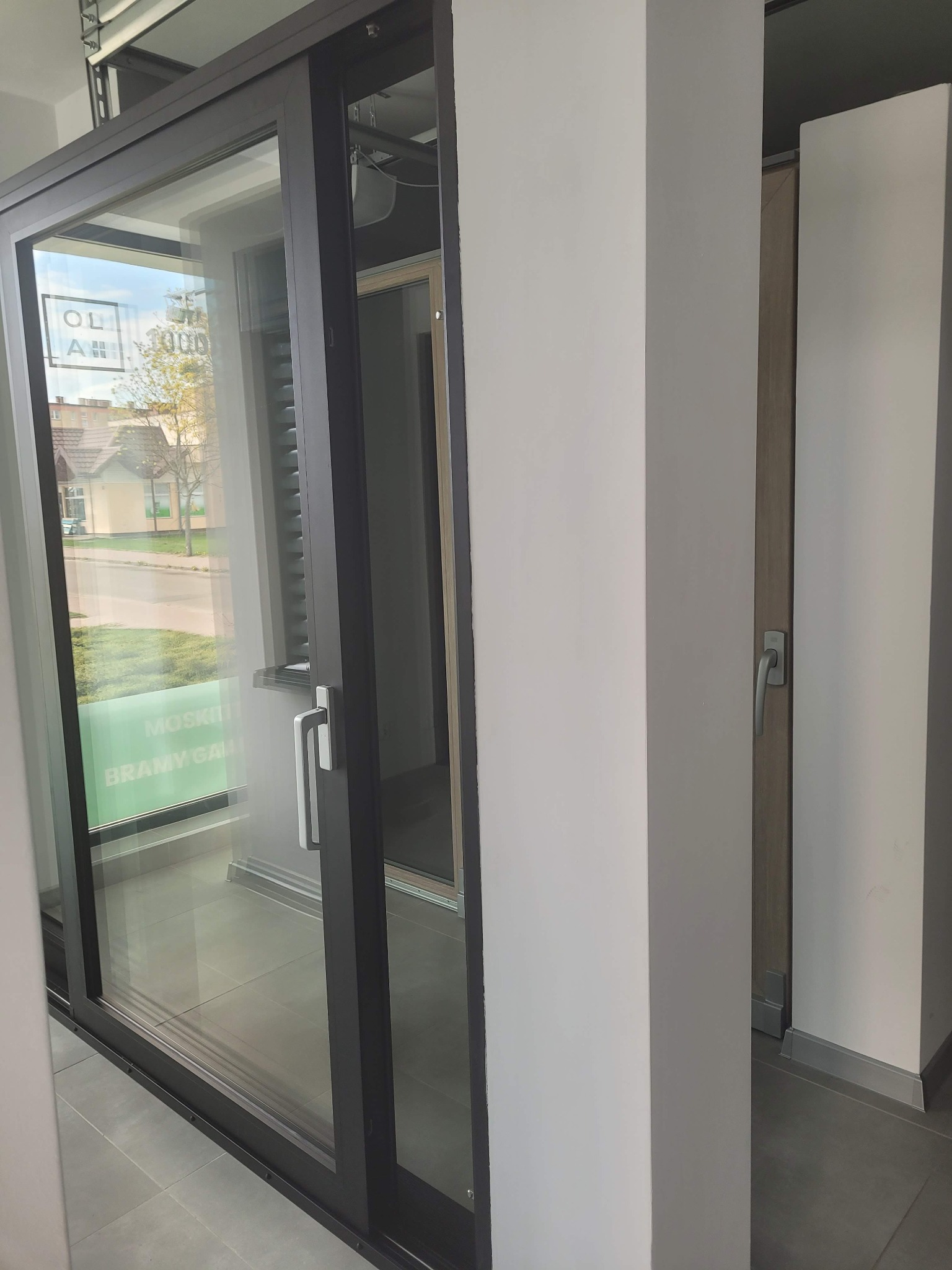 Ekspozycja w salonie sprzedaży OLA w Starogardzie Gdańskim: Czarna rama przesuwnych drzwi z widocznym uchwytem, odbicie ulicy i zieleni w szybie, oraz fragment ekspozycji innych drzwi w tle.