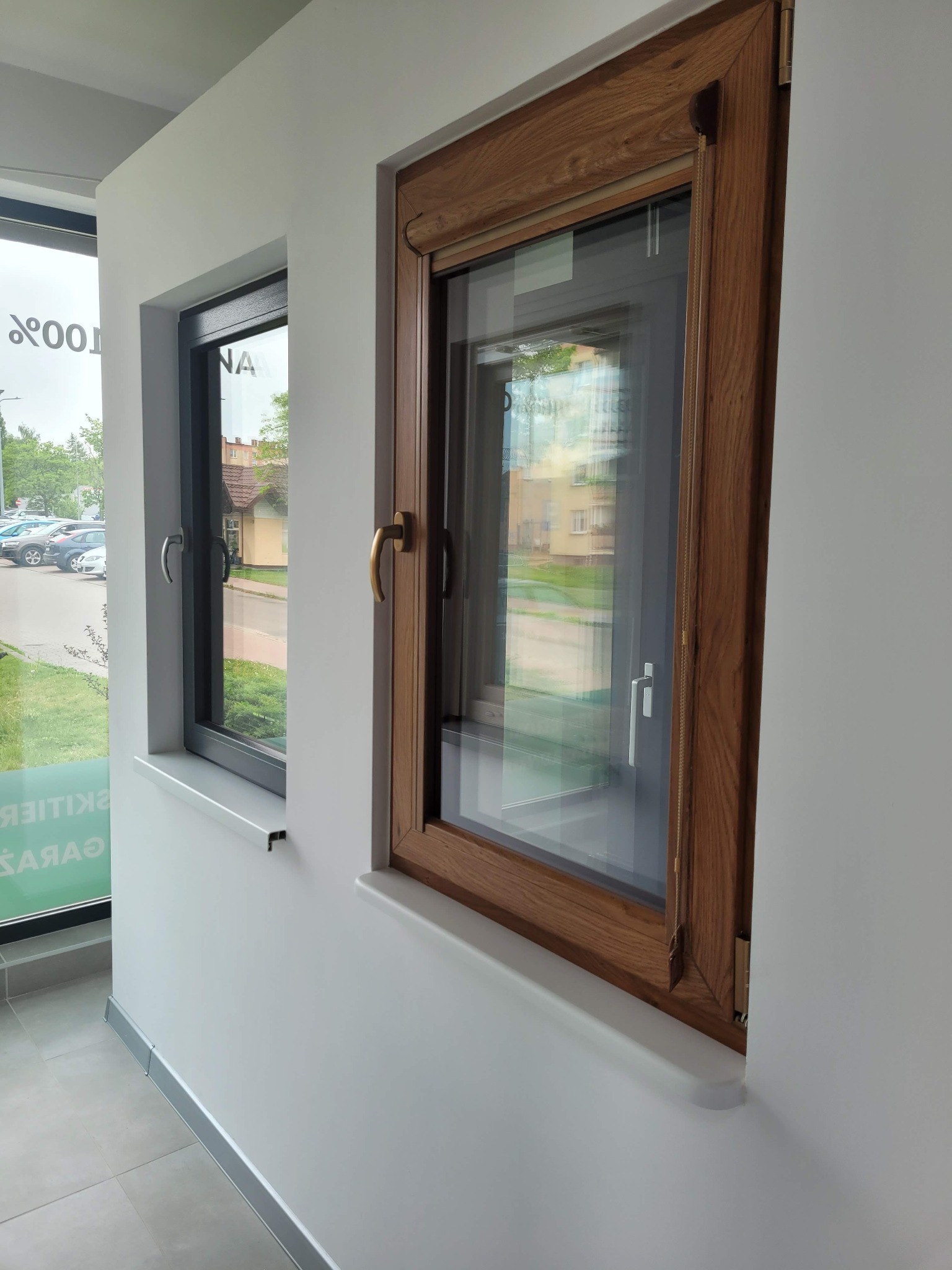 Prezentacja dwóch okien: szare okno z widokiem na parking i brązowe okno z wewnętrzną roletą sznurkową, oba zamontowane w białej ścianie.