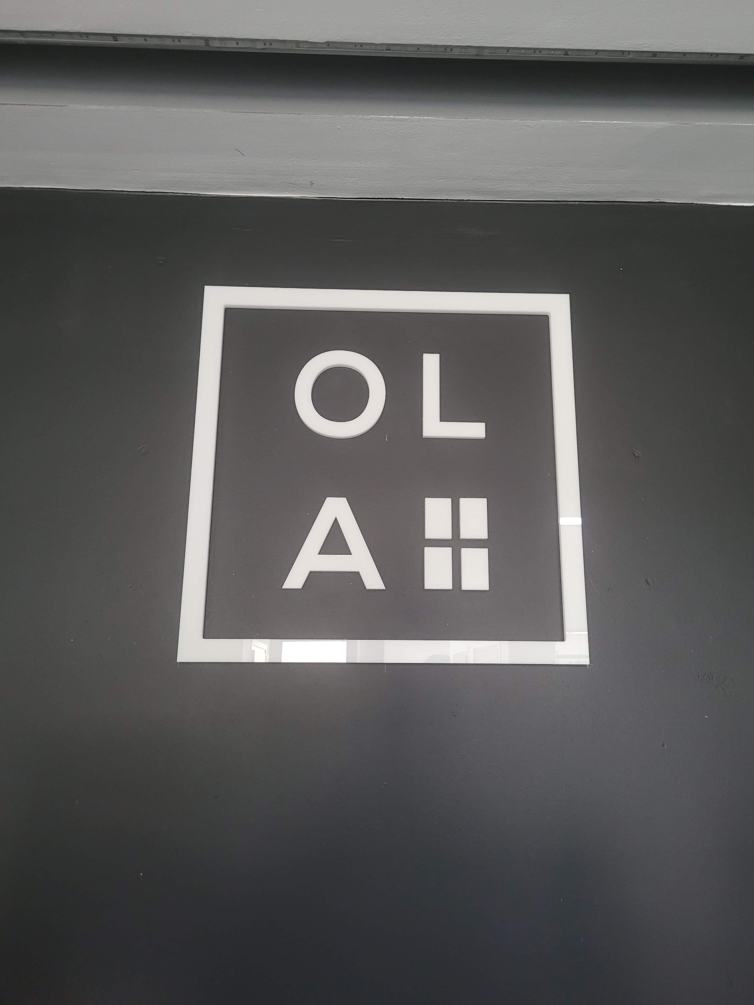 Biały, kwadratowy szyld z napisem 'OLA' i symbolem okna z czterema szybami na ciemnoszarej ścianie.