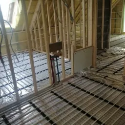 Montaż ogrzewania podłogowego w surowym wnętrzu poddasza, widoczne rury układane na folii z rastrem, drewniana konstrukcja szkieletowa ścian i ocieplenie poddasza.