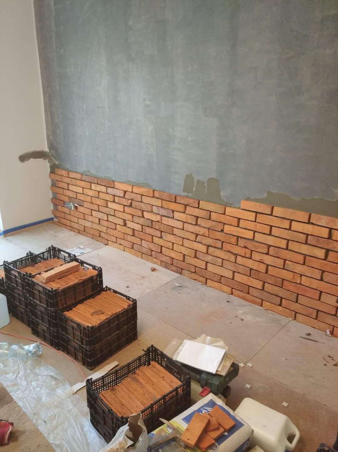Montaż cegieł dekoracyjnych na ścianie, widoczne kosze z cegłami, szara ściana w górnej części, etap wykończeniowy.