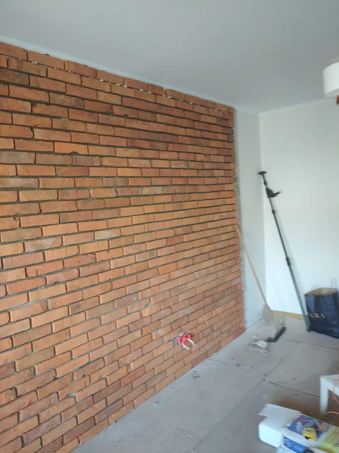Ściana z cegły klinkierowej w trakcie montażu z widocznymi przewodami elektrycznymi, narzędziami i workiem na podłodze.