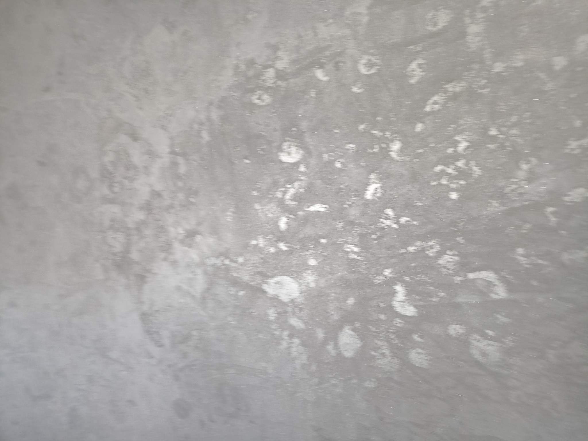 Szara ściana z nieregularnymi, jaśniejszymi plamami, przypominającymi efekt dekoracyjny lub niedoskonałe wykończenie.