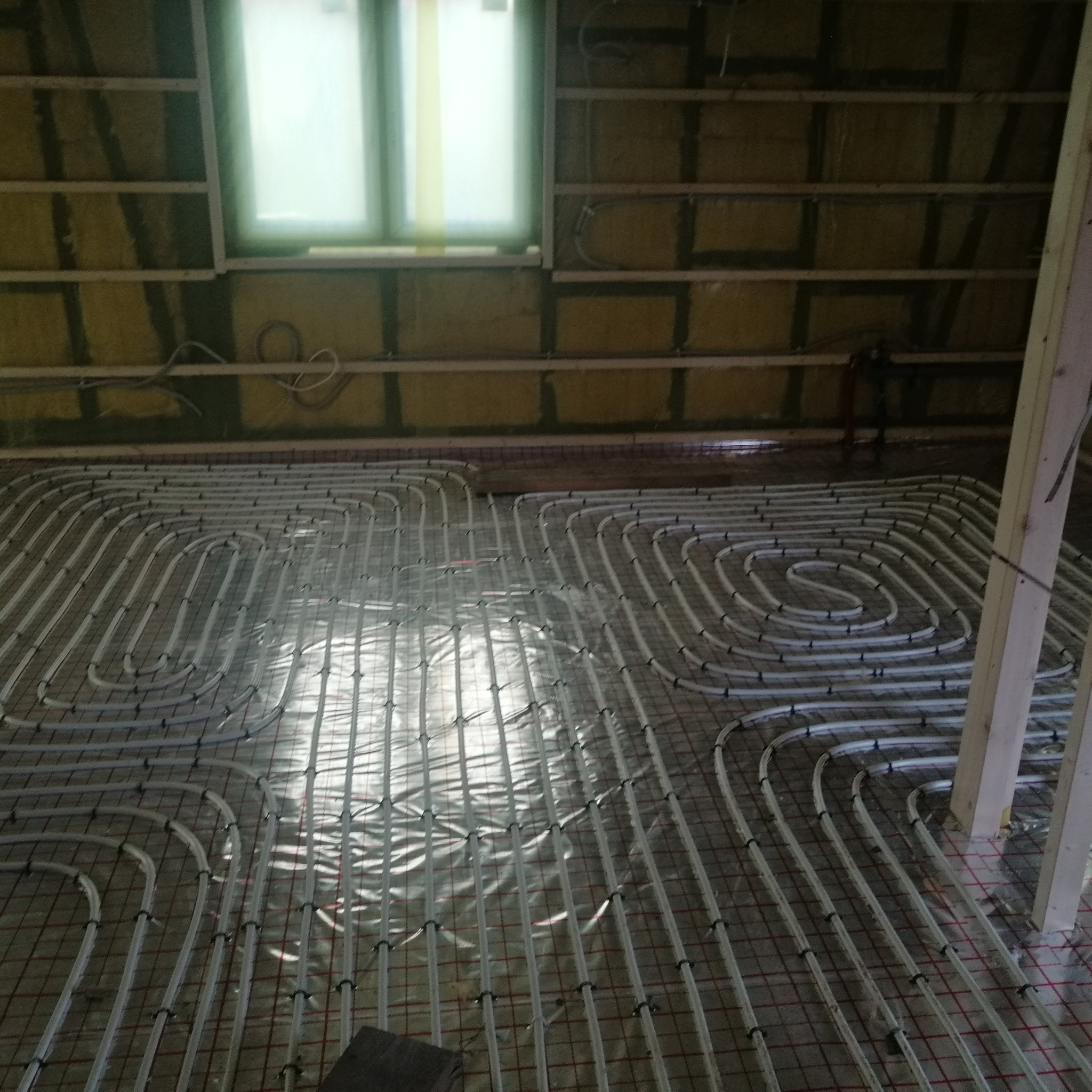 Instalacja ogrzewania podłogowego na siatce w pomieszczeniu z oknem, widoczne rury grzewcze ułożone w pętle i izolacja na ścianach poddasza.