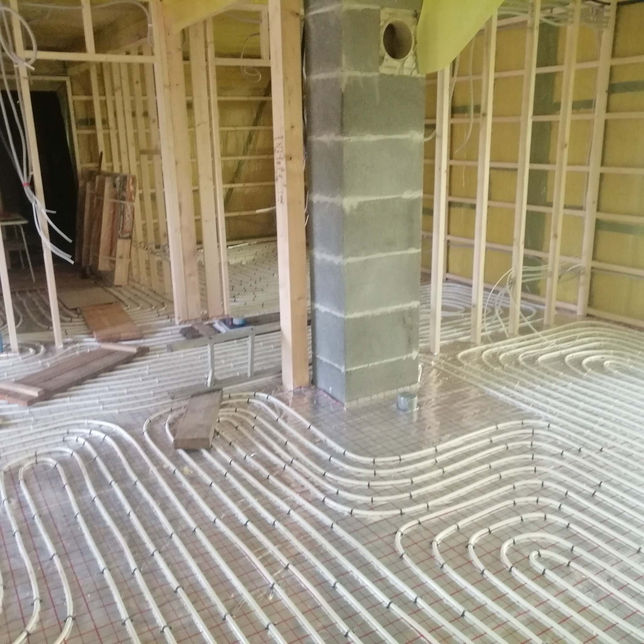 Montaż ogrzewania podłogowego: białe rurki ułożone na siatce w surowym wnętrzu z drewnianą konstrukcją ścian i betonowym kominem.