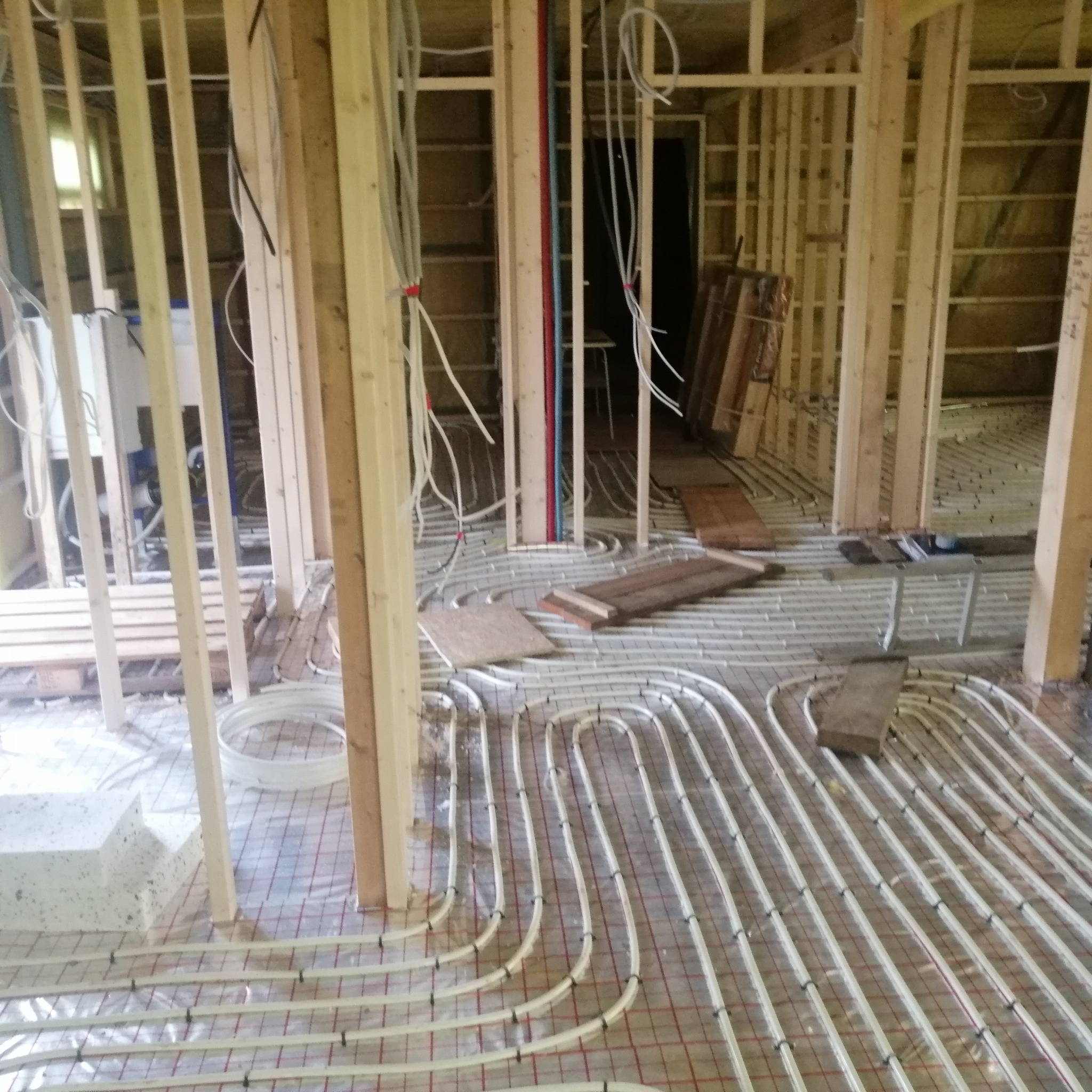 Instalacja ogrzewania podłogowego w trakcie budowy, widoczne rury grzewcze ułożone na siatce, drewniana konstrukcja szkieletowa ścian oraz instalacje elektryczne i wodno-kanalizacyjne.