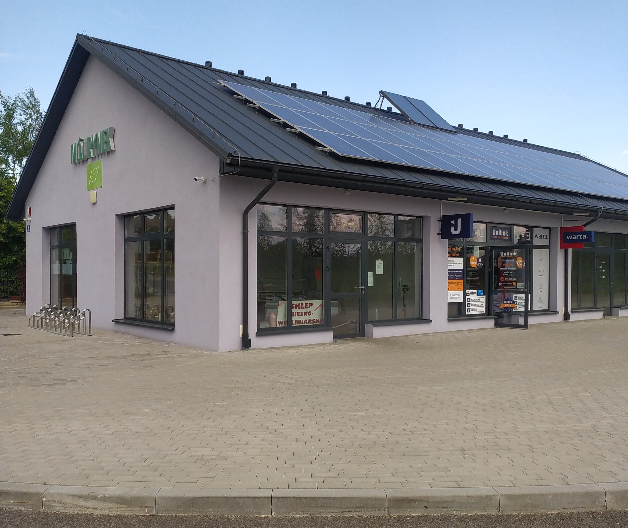 Budynek placówki partnerskiej Unilink S.A. z panelami słonecznymi na dachu i szyldami reklamowymi Warta.