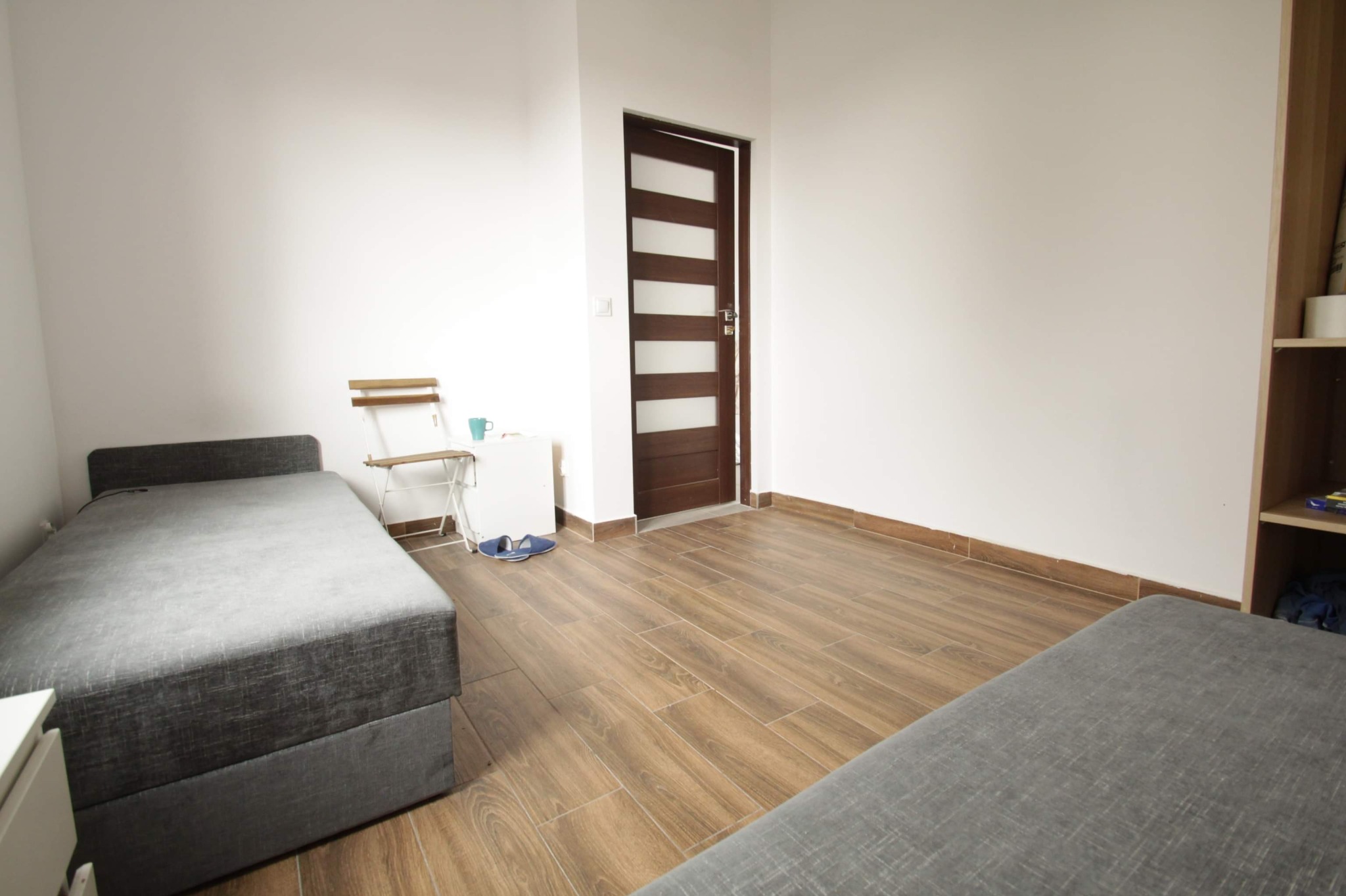 Jasne wnętrze pokoju z drewnopodobną podłogą, dwoma szarymi łóżkami, białymi ścianami, brązowymi drzwiami z przeszkleniami i krzesłem składanym przy ścianie.