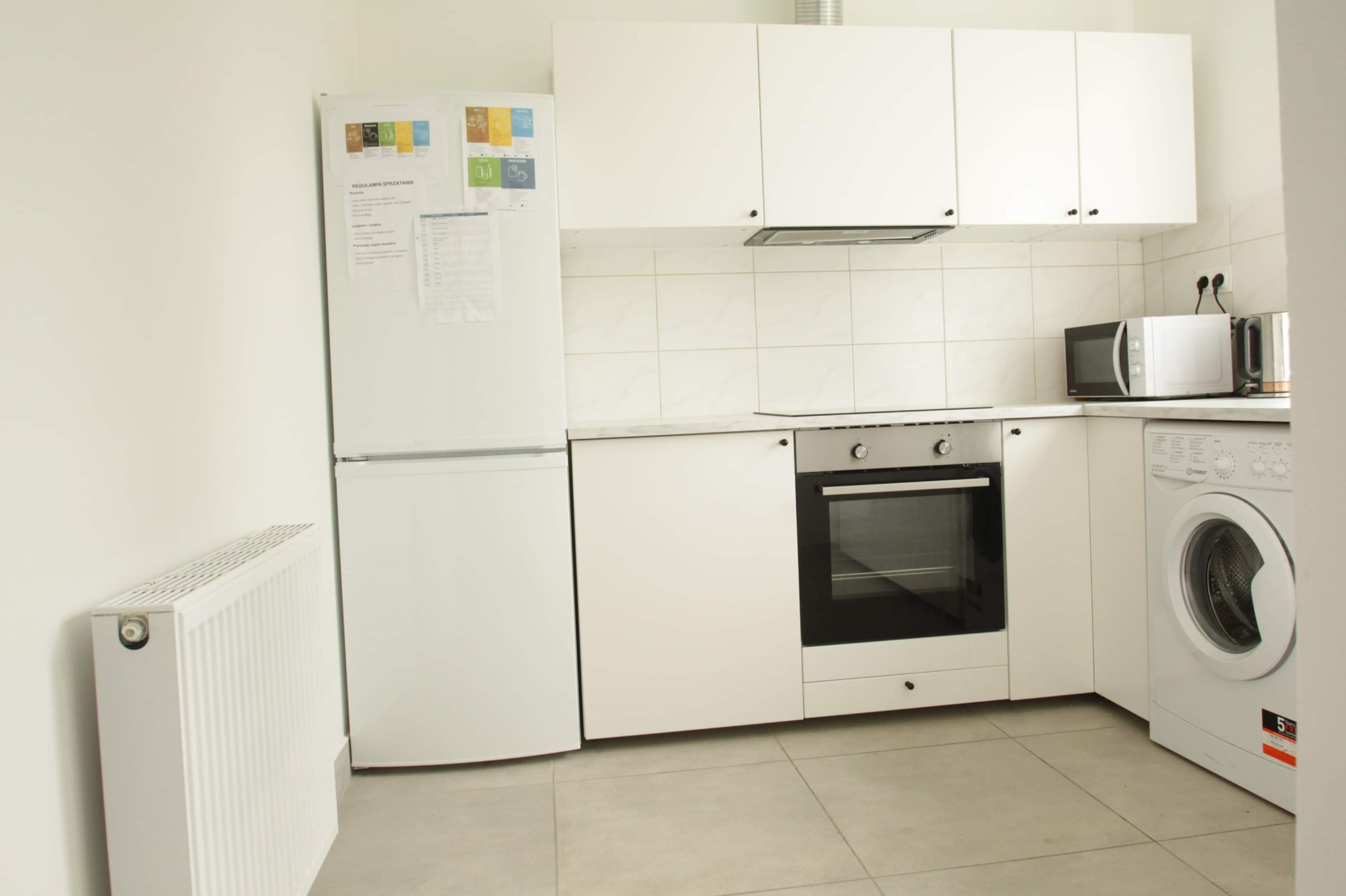 Jasna kuchnia w mieszkaniu w Warszawie po remoncie, z białymi meblami, sprzętami AGD i grzejnikiem, widok z niskiego kąta.