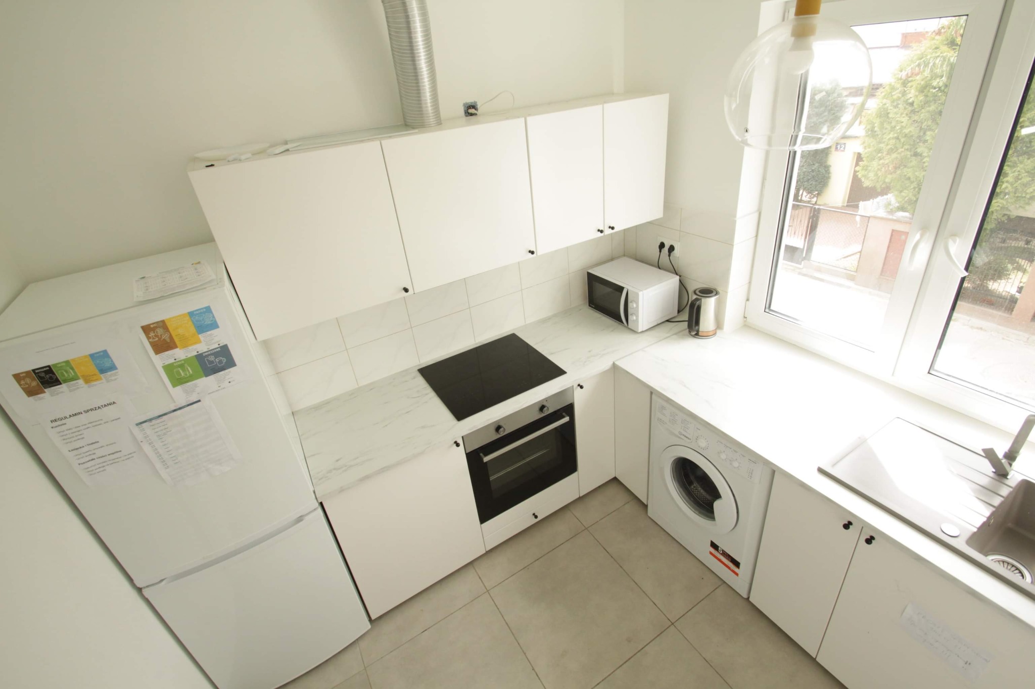Wnętrze jasnej kuchni po remoncie, z białymi szafkami, sprzętem AGD, marmurowym blatem i płytkami ściennymi, widziane z góry, z regulaminem sprzątania na lodówce.