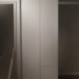 Prosta, biała szafa z czterema drzwiami, wbudowana we wnękę, widoczna w słabym oświetleniu, z fragmentem ościeżnicy po lewej stronie.