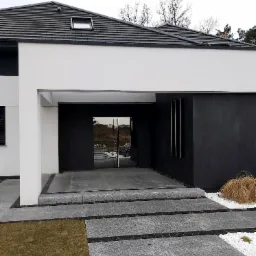 Nowoczesny dom z przeszklonymi drzwiami wejściowymi, elewacją w odcieniach bieli i grafitu, oraz ścieżką z szarej kostki granitowej z czarnymi wstawkami.