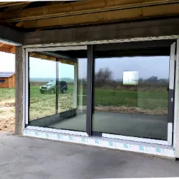 Duże przesuwne okno tarasowe z czarną ramą w surowym stanie budynku, widok na pole i budynek gospodarczy, na zewnątrz zaparkowany samochód.