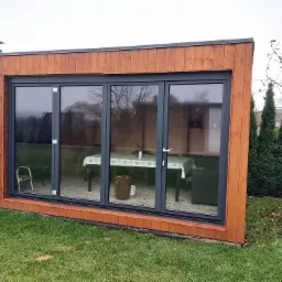 Nowoczesny budynek z drewnianą elewacją i dużymi, przesuwnymi oknami aluminiowymi w kolorze grafitowym, wychodzącymi na trawnik.