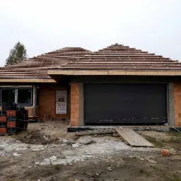 Budowa domu jednorodzinnego w trakcie: widać konstrukcję dachu z łatami, niezamontowane okno, czarną bramę garażową oraz paletę z dachówkami na placu budowy.