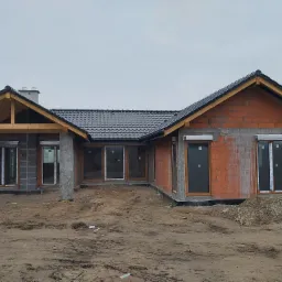 Surowy stan domu jednorodzinnego z zamontowanymi oknami z brązowymi ramami i szarymi roletami zewnętrznymi, widoczne niedokończone ściany z cegły i betonowe elementy konstrukcyjne.