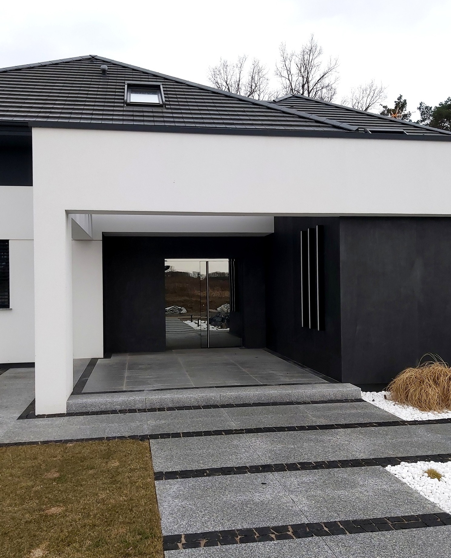 Nowoczesny dom z przeszklonymi drzwiami wejściowymi, elewacją w odcieniach bieli i grafitu, oraz ścieżką z szarej kostki granitowej z czarnymi wstawkami.