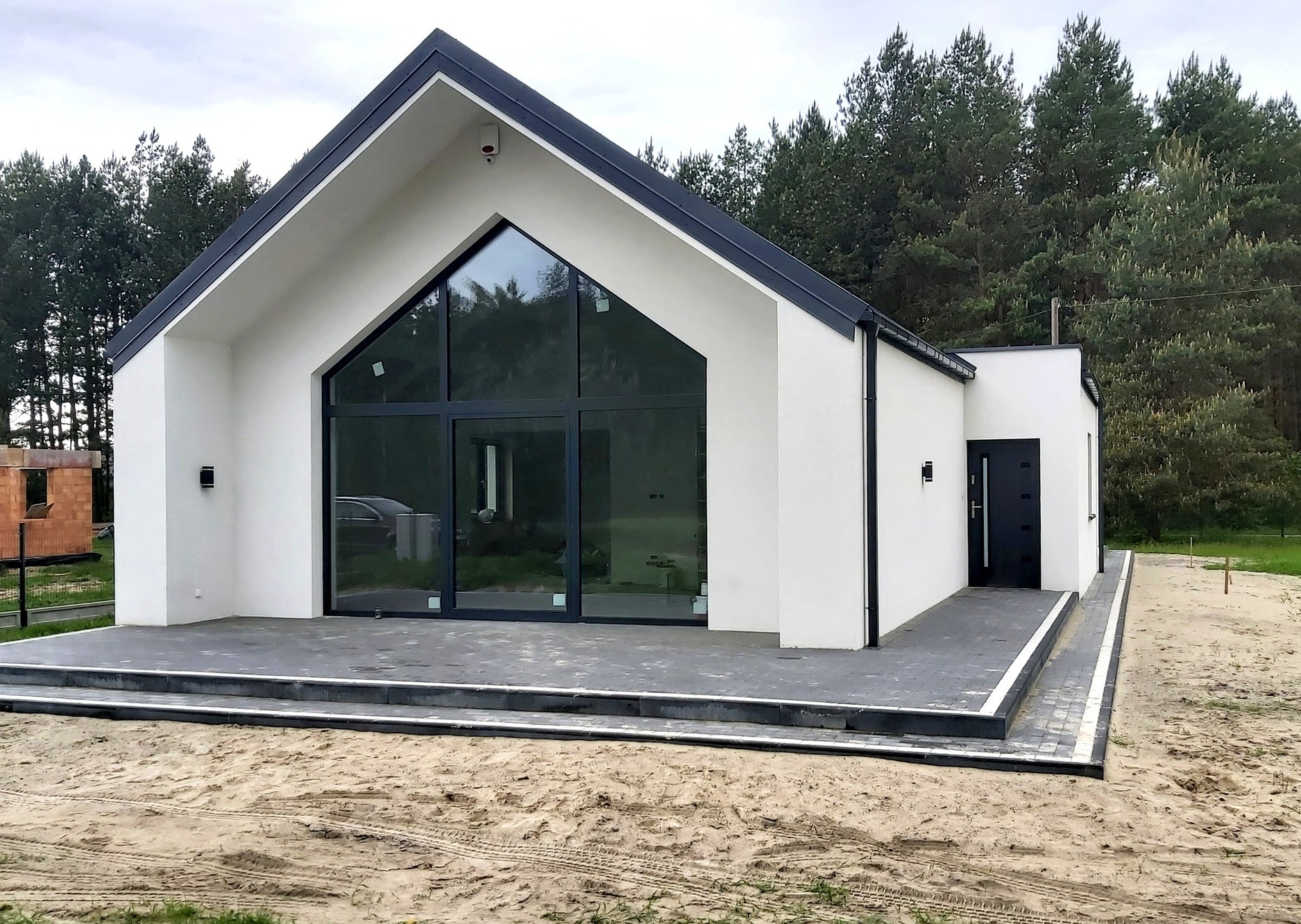 Nowoczesny dom jednorodzinny z dużym przeszkleniem w szczycie, antracytowa stolarka okienna i drzwiowa, minimalistyczna elewacja w kolorze białym, taras wyłożony szarą kostką, teren wokół domu...