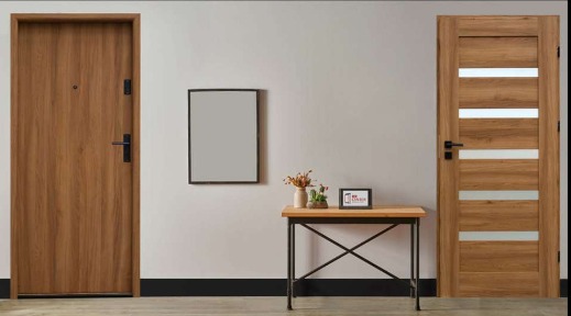 Dwa różne rodzaje drzwi wewnątrz budynku: brązowe drzwi antywłamaniowe z wizjerem po lewej i drewniane drzwi wewnętrzne z czterema poziomymi szybami po prawej stronie, na ścianie wisi czarna ramka,...