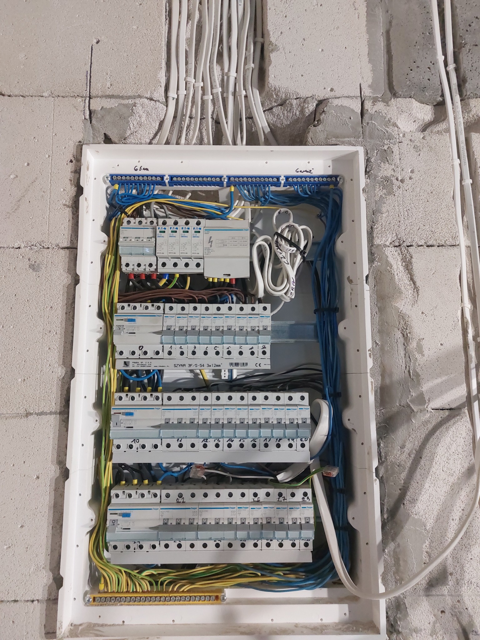 Rozdzielnica elektryczna z zainstalowanymi bezpiecznikami i przewodami w surowym wnętrzu, widoczne kable w białych peszlach w ścianie z betonu komórkowego.