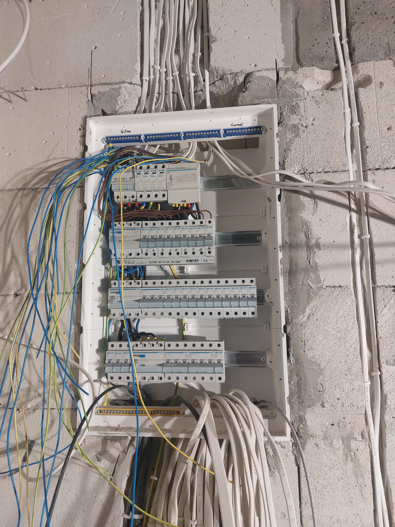 Rozdzielnica elektryczna w trakcie montażu w surowym wnętrzu, widoczne liczne kable w kolorach niebieskim, żółtym i zielonym, prowadzone do bezpieczników i listew zaciskowych. Ściana z betonu...