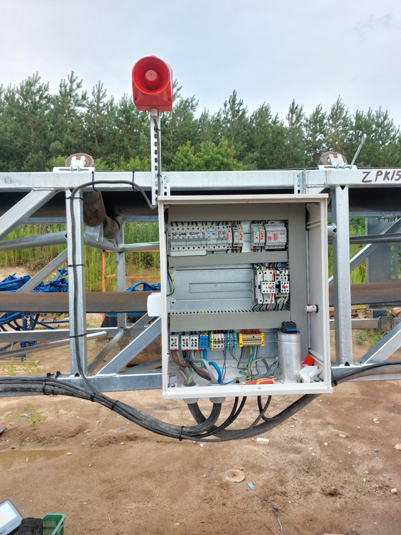 Otwarta szafa sterownicza z widocznymi elementami elektrycznymi i przewodami, zamontowana na metalowej konstrukcji na tle drzew i pochmurnego nieba. Czerwona syrena alarmowa na szczycie konstrukcji.