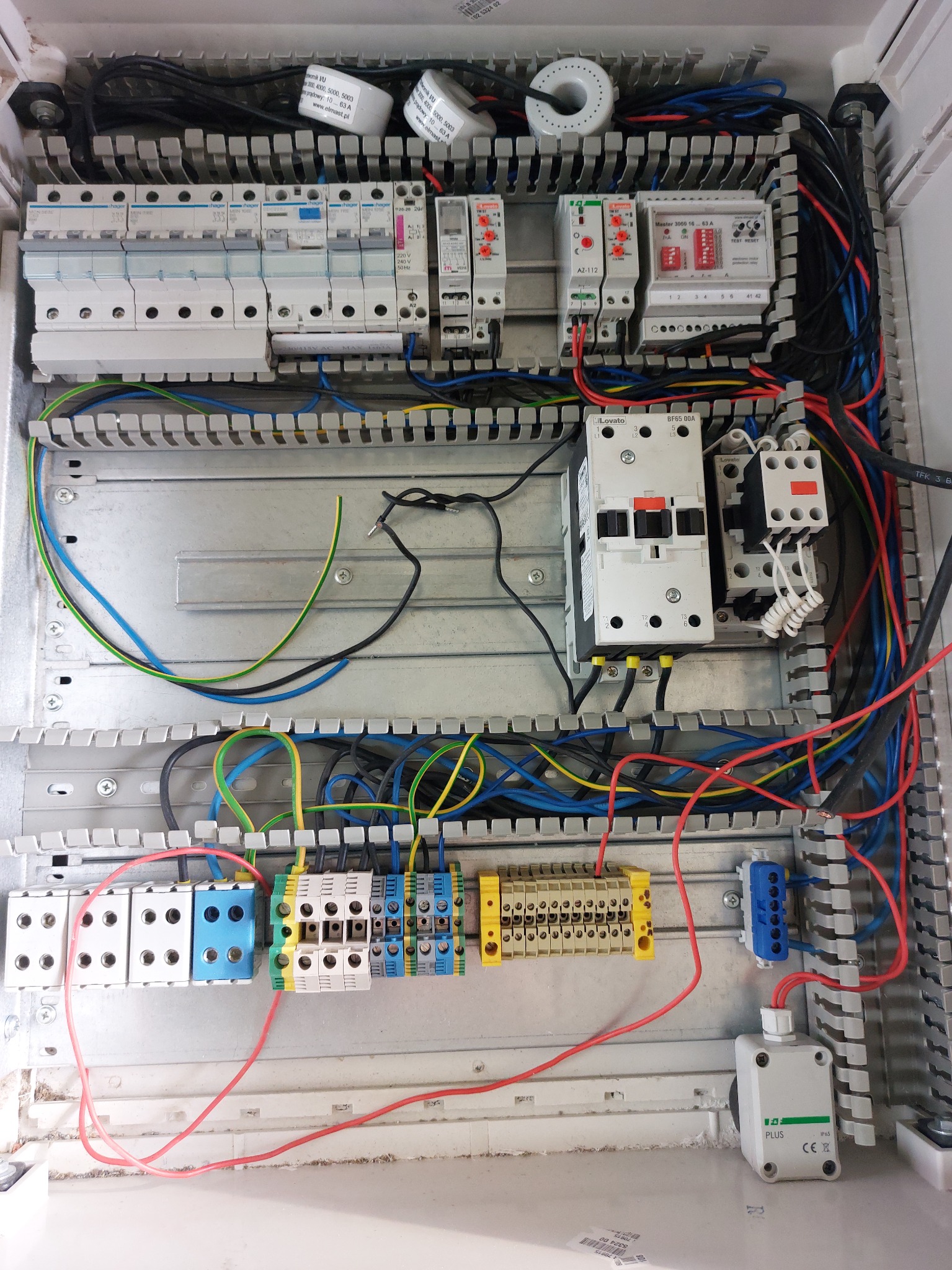 Otwarta skrzynka rozdzielcza z zainstalowanymi bezpiecznikami, przekaźnikami, stycznikami i listwami zaciskowymi, z widocznymi podłączeniami kablowymi w różnych kolorach.