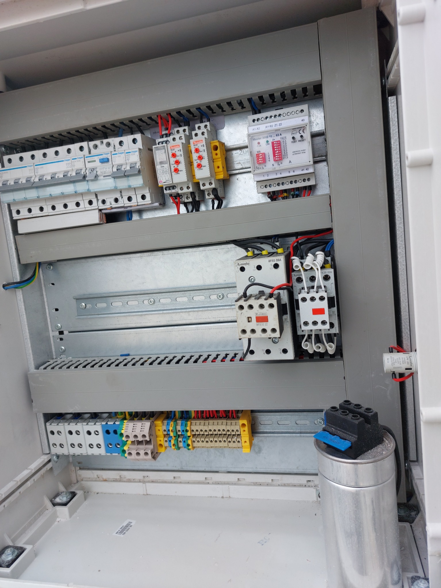 Otwarta skrzynka rozdzielcza z bezpiecznikami, przekaźnikami, stycznikami i listwami zaciskowymi, widoczne przewody w różnych kolorach, metalowa obudowa.