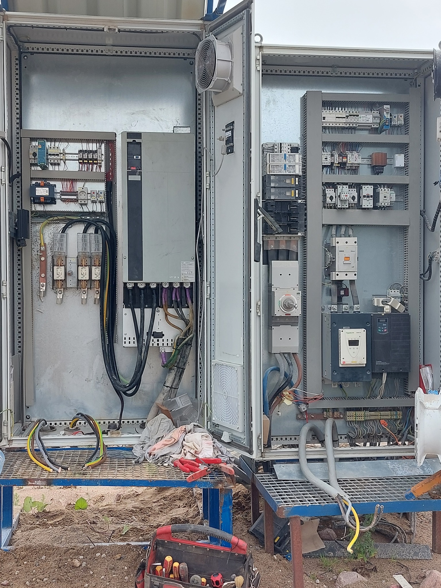 Otwarta szafa sterownicza z widocznymi elementami elektrycznymi, w tym rozdzielnicą, stycznikami i przewodami, na zewnątrz skrzynka z narzędziami i fragment podłoża z piaskiem.