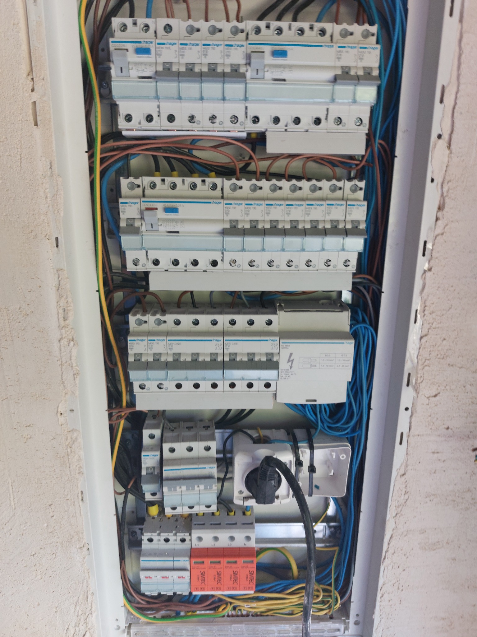 Rozdzielnica elektryczna Hager z zainstalowanymi bezpiecznikami i przewodami w kolorach niebieskim, brązowym, żółto-zielonym, zamontowana w ścianie w trakcie prac wykończeniowych.