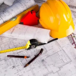 Żółty kask budowlany, poziomica, miara zwijana, dwa pióra kreślarskie i czerwona miniatura domu leżące na rozłożonych planach architektonicznych.