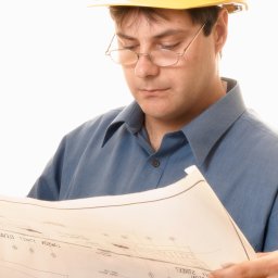 Odbiór mieszkania - Mężczyzna w okularach i żółtym kasku budowlanym analizuje rozłożony plan architektoniczny z odwróconymi napisami, ubrany w niebieską koszulę.