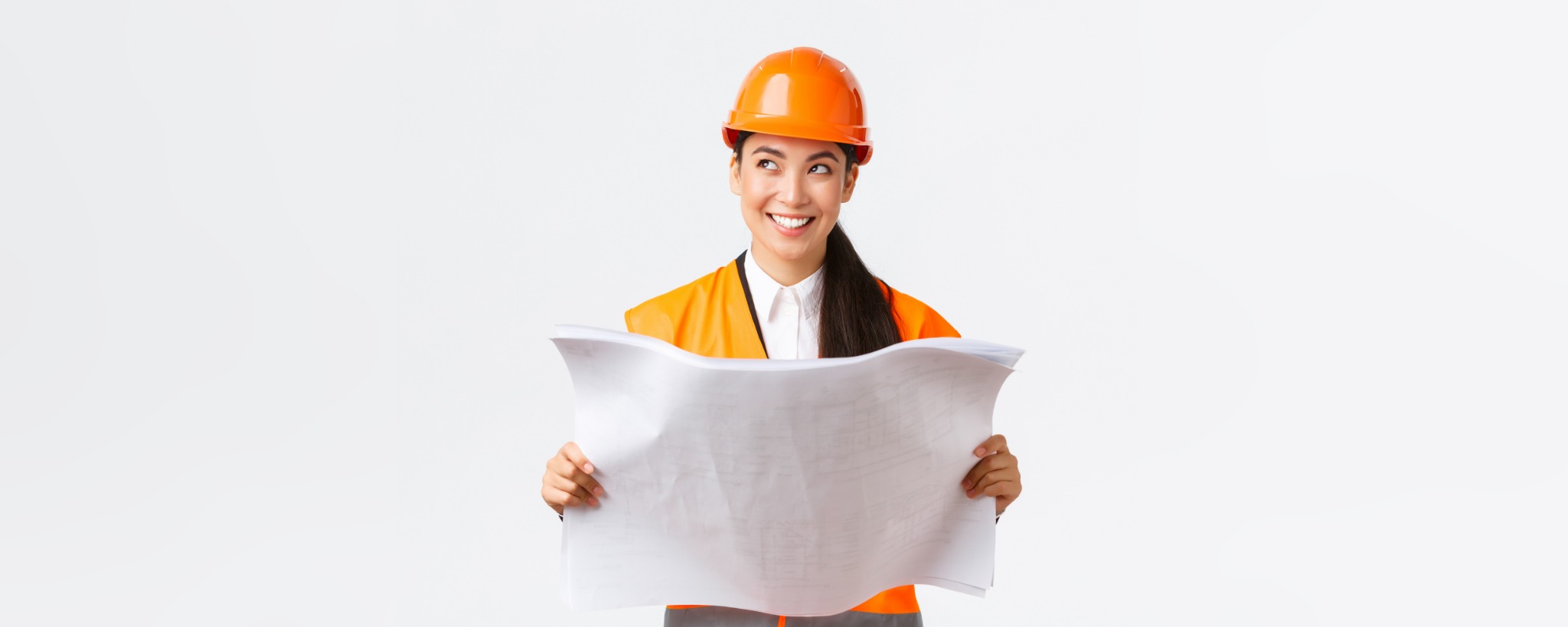 Uśmiechnięta kobieta w pomarańczowym kasku i kamizelce odblaskowej trzymająca rozłożony plan architektoniczny, spoglądająca w górę w lewo na białym tle.