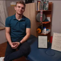 Młody rehabilitant z Fałkowa siedzi na niebieskim stole do masażu obok swoich dyplomów, w tle regał z książkami i przyborami do ćwiczeń.