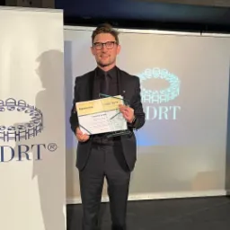 Mężczyzna w czarnym garniturze i okularach trzyma certyfikat i szklaną statuetkę z napisem MDRT 2022, na tle logo MDRT.