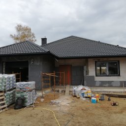 Domy Energo - Oszczędne CANADA SYSTEM - Budowa domu w trakcie ocieplania: ściany z szarej izolacji, palety z workami materiałów budowlanych, rusztowanie i narzędzia na placu budowy, widoczny fragment dachu z czarną dachówką.