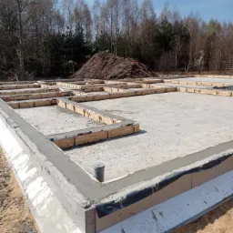 Płyta fundamentowa z izolacją obwodową i przygotowanymi szalunkami pod ściany, widok na plac budowy z hałdą ziemi w tle i drzewami na horyzoncie.