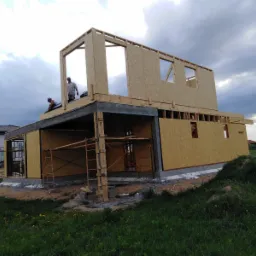 Wznoszenie dwukondygnacyjnego domu z płyt OSB na betonowym fundamencie; widoczne rusztowania i dwóch pracowników na górze, pochmurne niebo w tle.