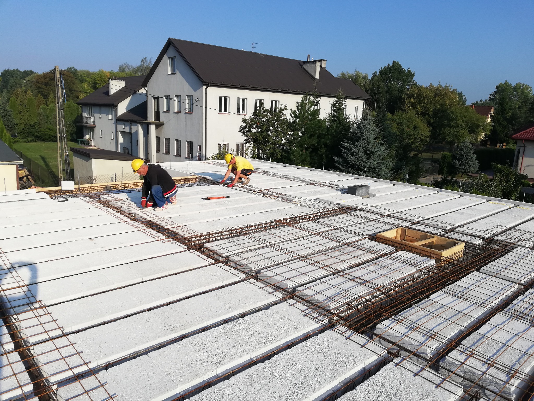 Dwóch robotników w kaskach układa zbrojenie na stropie z prefabrykatów betonowych, widok z góry na budowę domu jednorodzinnego z otoczeniem zieleni i innymi budynkami.
