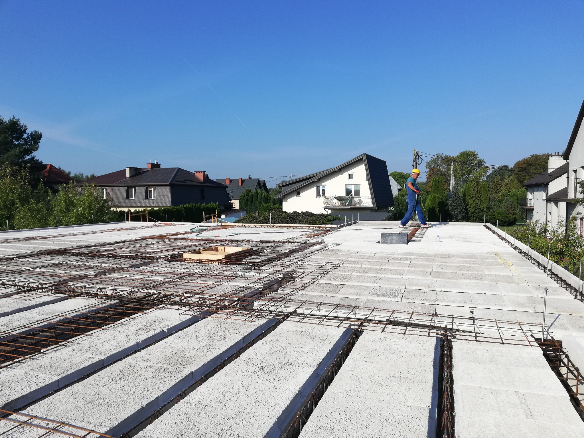 Prace na budowie: robotnik w niebieskim kombinezonie i żółtym kasku stoi na stropie Teriva, widoczne zbrojenie i bloczki betonowe, w tle zabudowa jednorodzinna i błękitne niebo.