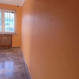 Usługi Malarskie Warszawa Robert Gawlik - Pokój z pomarańczową ścianą, widoczne ślady po wcześniejszych mocowaniach na ścianie, okno z ozdobnymi szprosami i stary grzejnik.