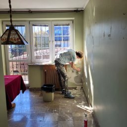 Usługi Malarskie Warszawa Robert Gawlik - Remont pokoju: kobieta szpachluje ścianę przy oknie, widoczne ślady po usuniętej farbie, narzędzia i wiadro na podłodze.