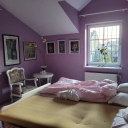 Usługi Malarskie Warszawa Robert Gawlik - Fioletowy pokój na poddaszu z łóżkiem bez pościeli, ozdobnym krzesłem i obrazami na ścianie, oświetlony popołudniowym światłem z okna.